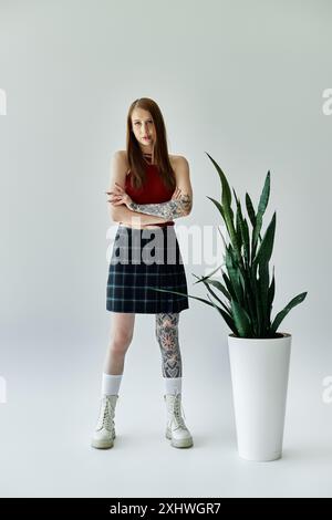 Une jeune femme avec des tatouages sur les bras et les jambes se tient dans une jupe à carreaux et un débardeur devant un fond gris, avec une grande plante à sa droite. Banque D'Images