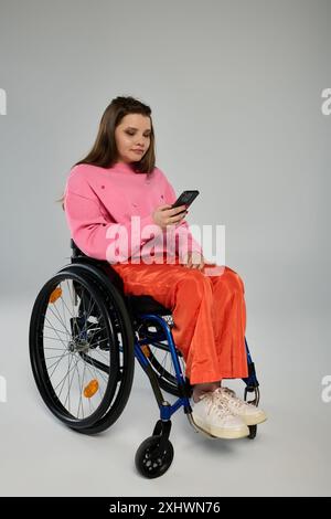 Une jeune femme brune portant des vêtements décontractés est assise dans un fauteuil roulant et utilise son smartphone. Banque D'Images