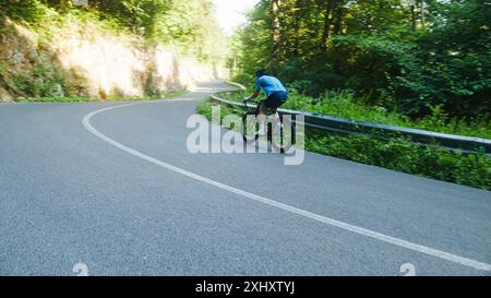 Cycliste de course automobile masculin dans un maillot de sport bleu, avec un casque et des lunettes, en montant une colline à travers la forêt. Banque D'Images