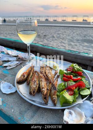 Assiette de poisson grillé avec salade fraîche et un verre de vin blanc sur une table en bord de plage au coucher du soleil, parfait pour une soirée relaxante. Banque D'Images