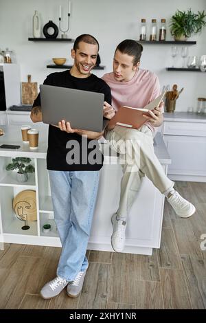 Un jeune couple gay profite d'un après-midi agréable dans leur cuisine moderne, l'un naviguant sur un ordinateur portable tandis que l'autre lit un livre. Banque D'Images