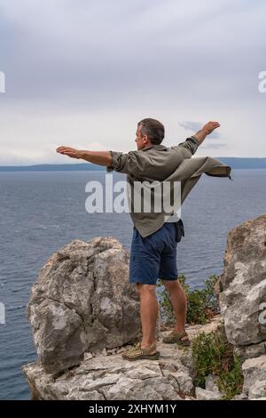 Un homme d'âge mûr profitant de son voyage. Se trouve au bord d'une falaise et regarde le paysage marin. Il écarta les bras sur les côtés. La chemise bat en t Banque D'Images