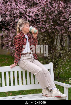 Vue de côté d'une jolie jeune femme buvant calmement du café dans une tasse. Une fille aux cheveux blonds est assise sur un banc dans le parc, profitant de la vie, une courte pause Banque D'Images