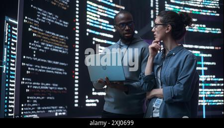 Portrait de deux développeurs divers utilisant un ordinateur portable, discutant des lignes de code qui apparaissent sur les grands écrans qui les entourent. Programmeurs masculins et féminins créer un logiciel ensemble et corriger des bugs Banque D'Images