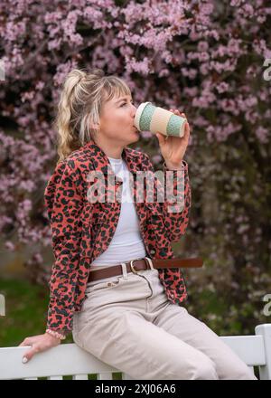 Une jolie fille blonde caucasienne est assise sur un banc et aime boire du café. Belle femme passe son temps libre. Concept de style de vie, pause. Banque D'Images