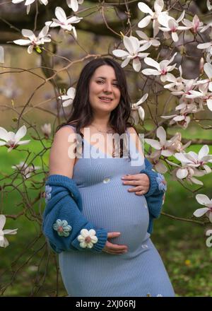 Portrait d'une jolie femme enceinte dans le parc, vêtue d'une robe bleue parmi les fleurs de magnoli. une femme tient doucement son ventre enceinte. Grossesse. Attendez Banque D'Images