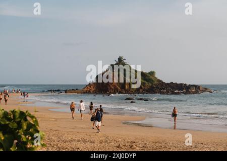 Les gens marchent le long de Mirissa Beach au Sri Lanka, avec un îlot rocheux et couvert de végétation Parrot Rock près de la rive. Banque D'Images