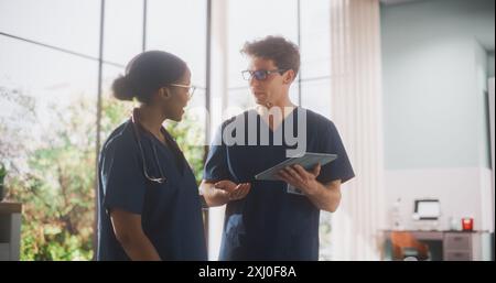 Deux jeunes médecins debout dans un établissement médical de l'hôpital, parler et partager des informations sur un ordinateur tablette. Hôpital moderne avec divers professionnels de la santé multiethniques Banque D'Images