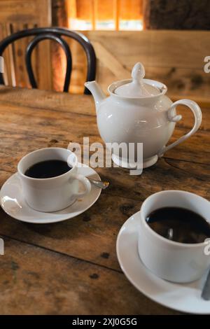 Une table en bois avec deux tasses de café et une théière blanche. Le cadre est dans un petit restaurant rural et agolden soleil illuminant la scène. Banque D'Images