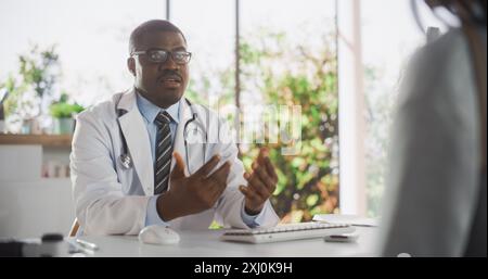 Docteur noir parlant à un patient anonyme lors d'une consultation en clinique de santé. Médecin en White Lab Coat suggérant un traitement au visiteur dans le bureau de l'hôpital moderne Banque D'Images