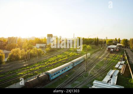 Railyard au coucher du soleil. Triage à moitié vide avec peu de wagons qui peuvent sembler abandonnés. Soleil bas et haute herbe verte dans la zone industrielle. Banque D'Images