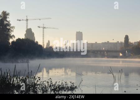 Aube brumeuse dans une ville. Brouillard sur l'eau du canal fluvial. Pont et silhouettes de bâtiments rétroéclairés par le soleil du matin. Chantier de construction avec grues à proximité. Banque D'Images