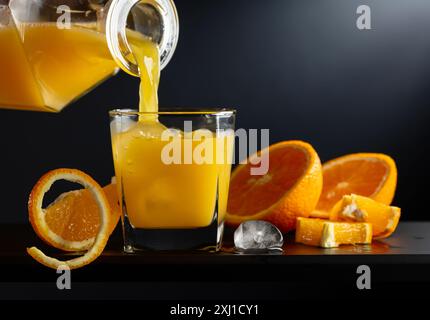 Verre de jus d'orange avec de la glace sur un fond noir. Le jus est versé d'une carafe sur un verre avec de la glace. Boisson rafraîchissante avec des tranches de fruits. Banque D'Images