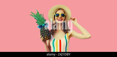 Vacances d'été, tourisme, jeune femme joyeuse heureuse avec des fruits d'ananas dans le chapeau de touriste, fille joyeuse posant sur fond de studio rose Banque D'Images