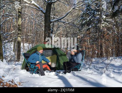 Un homme et une femme sont assis sur des chaises dans une forêt enneigée près d'une tente. Préparation de thé. Vie active. Joie et contentement dans la vie. Air givré, brûlant Banque D'Images