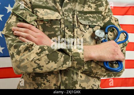 Homme en uniforme militaire avec les bras croisés et stéthoscope contre le drapeau américain, gros plan. Concept de soins de santé Banque D'Images