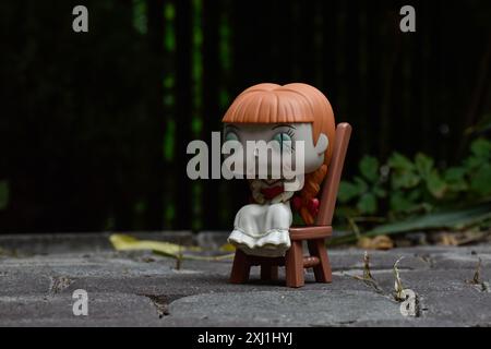 Funko Pop figurine d'action de poupée hantée Annabelle assise sur la chaise des films d'horreur conjurant. Cour sombre, clôture noire, ombres, vert sombre. Banque D'Images