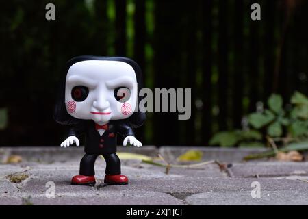 Funko Pop figurine d'action de Billy la marionnette du populaire film d'horreur Slasher Saw. Cour sombre, clôture noire, ombres, fond vert sombre. Banque D'Images