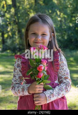 Une petite fille mignonne dans une robe rose tient des roses dans ses mains et les regarde gaiement. Banque D'Images