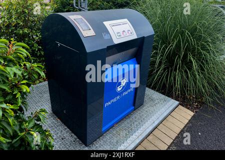 Bac de recyclage de papier gris avec un panneau bleu marqué papier, Pappe est assis sur une plate-forme en béton dans un cadre urbain. Du feuillage vert entoure le bac. Banque D'Images