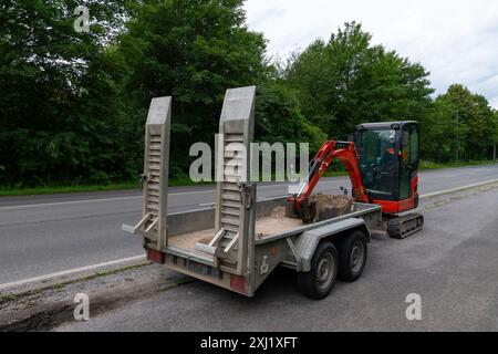 Une pelle rouge est chargée sur une remorque sur le bord d'une route, prête pour le transport. La remorque est dotée de deux rampes abaissées et le godet de la pelle repose sur le sol. Banque D'Images