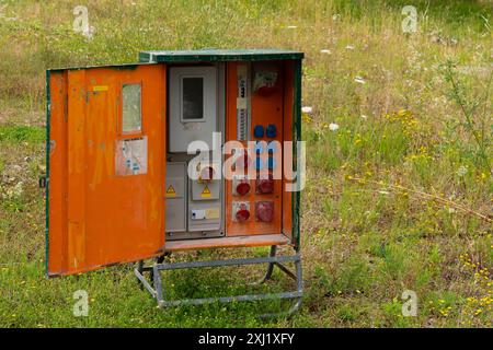 Une vieille boîte électrique orange avec une porte métallique ouverte, révélant une variété de prises électriques, des interrupteurs et un compteur. La boîte est située dans un champ avec de l'herbe verte et des fleurs sauvages jaunes. Banque D'Images