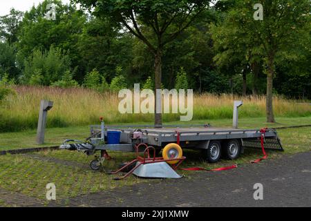 Une remorque à plateau est stationnée dans une zone herbeuse à côté d'une rangée d'arbres. La remorque est vide et une cale de roue est en place. Il y a une brouette et un seau bleu sur le sol près de la remorque. Banque D'Images