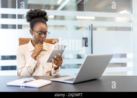 Femme d'affaires concentrée tenant une tablette numérique tout en travaillant au bureau avec ordinateur portable et livre dans un cadre de bureau moderne. Environnement professionnel, technologie, concentration et concept de productivité. Banque D'Images