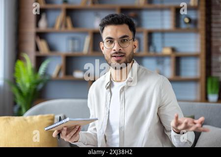 Portrait en gros plan d'un jeune musulman en lunettes et chemise qui est à la maison, tenant un stylo et un cahier et agitant ses bras de manière inexplicable à la caméra. Banque D'Images