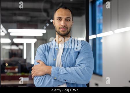 Portrait d'un homme souriant et confiant debout dans une chemise dans un bureau moderne, croisant ses bras sur sa poitrine et regardant la caméra. Banque D'Images