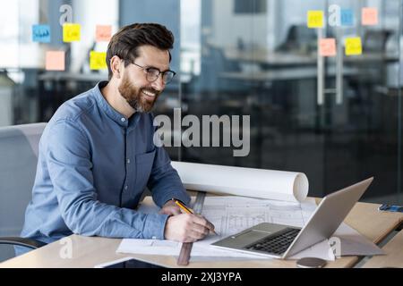 Un jeune homme souriant architecte, ingénieur et développeur travaille dans le bureau selon des dessins, s'assoit à un bureau avec des papiers, parle lors d'un appel vidéo. Banque D'Images