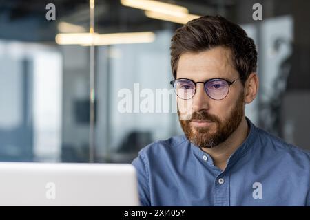 Jeune programmeur et développeur assis dans un bureau moderne portant des lunettes et une chemise et travaillant sur un ordinateur portable en ligne. Banque D'Images