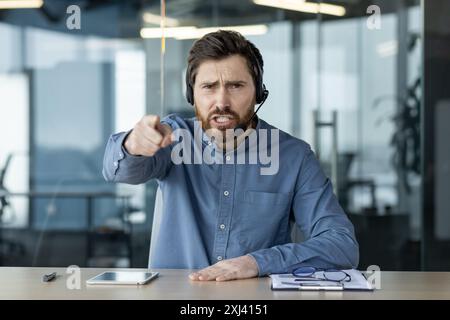 Portrait d'un jeune homme dans un casque assis dans le bureau à la table, parlant émotionnellement en ligne, criant et pointant vers la caméra. Banque D'Images