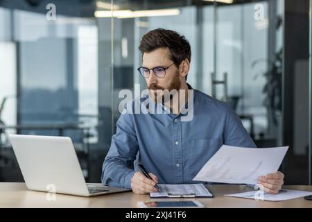 Financier sérieux dans le bureau moderne travaillant sur la paperasse, analysant les données sur ordinateur portable. Jeune comptable avec barbe et lunettes axé sur les documents d'affaires. Environnement professionnel sur le lieu de travail Banque D'Images