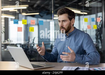 Un jeune homme d'affaires sérieux est assis dans le bureau à son bureau et semble choqué par l'écran de l'ordinateur portable. Banque D'Images