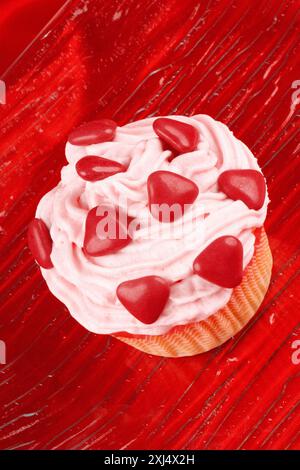 Cupcake fantaisie de la Saint-Valentin décoré de petits coeurs en chocolat enrobés de sucre rouge sur un plat en verre transparent, sur fond rouge. Sélectif Banque D'Images