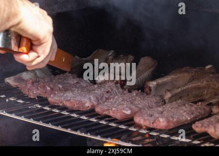 Griller en action : un chef déverse habilement des viandes grésillantes sur un barbecue fumé, capturant l'essence de la cuisine en extérieur et culinaire estivale Banque D'Images