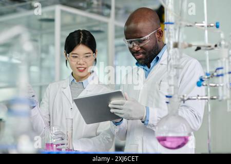 Deux jeunes chimistes interculturels regardant l'écran de tablette tenu par un scientifique afro-américain tout en effectuant un nouvel essai Banque D'Images