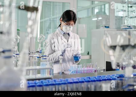 Jeune chercheuse en respirateur de protection debout par le lieu de travail dans le laboratoire et mélangeant deux substances chimiques liquides Banque D'Images