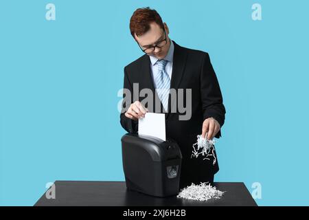 Beau jeune homme détruisant la feuille de papier dans la déchiqueteuse sur fond bleu Banque D'Images