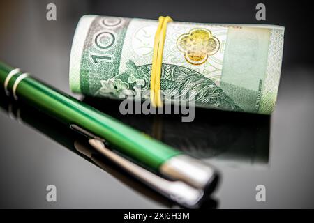 Monnaie polonaise, 100 billets de banque Zloty roulés et sécurisés avec un élastique jaune, mis en évidence à côté d'un stylo vert élégant sur un fond brillant, emphasi Banque D'Images
