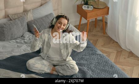 Vue de dessus heureuse femme caucasienne assise lit écouteurs smartphone téléphone mobile gadget technologie électronique écouter de la musique chantant fun énergique Banque D'Images
