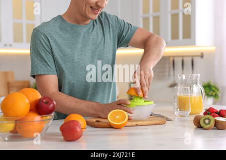 Homme pressant l'orange fraîche avec presse-agrumes à la table en marbre blanc dans la cuisine, gros plan Banque D'Images