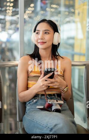 Une belle touriste asiatique avec des écouteurs et un smartphone sur sa main, assise à un siège d'attente dans le terminal de l'aéroport, regardant loin de th Banque D'Images