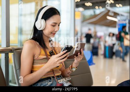 Une belle jeune touriste asiatique tient son smartphone et son passeport, vérifiant ses informations de vol sur son téléphone tout en étant assise à une attente Banque D'Images