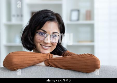Une jeune femme avec des lunettes sourit chaleureusement, reposant ses bras sur un canapé dans un cadre confortable à la maison. Banque D'Images