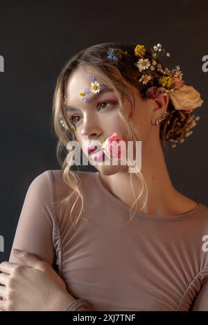 Jeune femme aux longs cheveux blonds ornés de fleurs regarde sur le côté, son visage également décoré de fleurs délicates. Banque D'Images