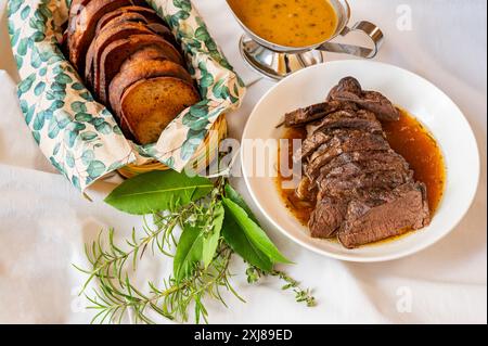 Longe de venaison cuite au four, sauce orange sauvage dans une casserole et tranches de pain grillé dans un bol sur une serviette à feuilles, bouquet d'herbes sur une nappe blanche, gros plan. Banque D'Images