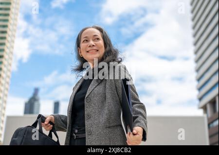 Une femme d'affaires senior asiatique confiante et heureuse se tient dans la grande ville urbaine, tenant un dossier de documents, regardant le ciel et les bâtiments avec un rêve, Banque D'Images