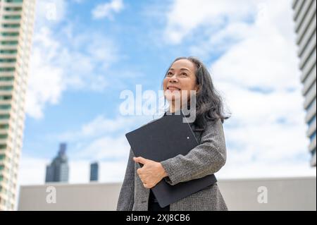 Une femme d'affaires senior asiatique confiante et heureuse se tient dans la grande ville urbaine, tenant un dossier de documents, regardant le ciel et les bâtiments avec un rêve, Banque D'Images